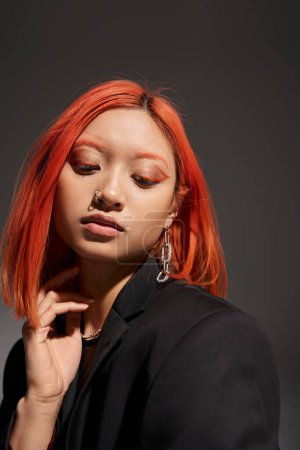 retrato de mujer joven asiática con pelo rojo, piercing en la nariz y maquillaje de ojos posando sobre gris