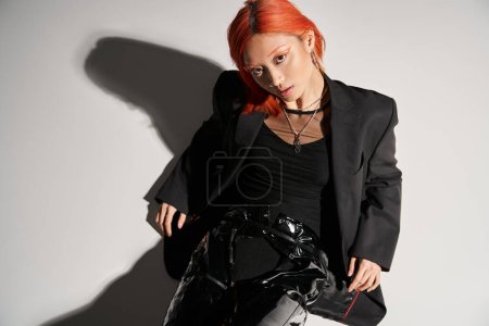 top view of asian woman with red hair posing in bold attire on grey background, black latex