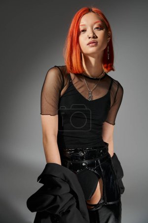 fashionable asian woman with red hair and nose piercing posing in trendy attire on grey background