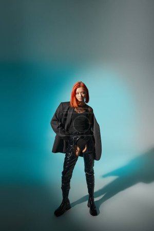 stylish asian woman with dyed hair posing in oversized blazer and latex boots on blue background
