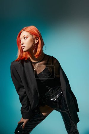 modelo asiático con pelo rojo y piercing posando en chaqueta de gran tamaño y látex negro sobre fondo azul