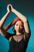 young asian woman posing with raised hands in black transparent blouse on blue backdrop, edgy look Poster #673633286