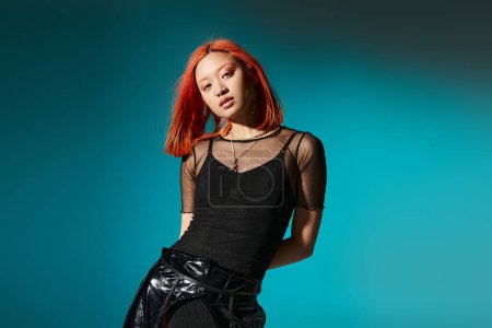 sassy asian woman with piercing posing in black latex pants and transparent blouse on blue backdrop