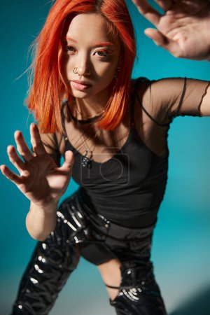 asiatisches Model mit Piercing und rot gefärbten Haaren gestikuliert und blickt in die Kamera auf blauem Hintergrund