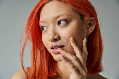 belleza asiática, mujer joven con el pelo rojo y maquillaje natural mirando hacia otro lado y tocando mejilla
