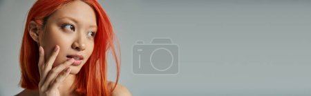 belleza asiática, mujer joven con el pelo rojo y maquillaje natural mirando hacia otro lado y tocando mejilla, bandera