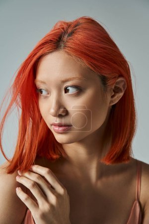 élégante et jeune femme asiatique aux cheveux roux et maquillage naturel regardant loin sur fond gris