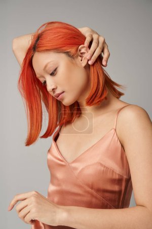 portrait of young asian woman with perfect skin adjusting red hair on grey backdrop, feminine grace