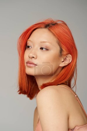Porträt einer anspruchsvollen jungen Asiatin mit perfekter Haut und roten Haaren, die auf grauem Hintergrund posiert