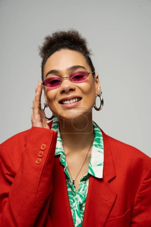 mujer afroamericana feliz y elegante en gafas de sol de moda y pendientes de aro sobre fondo gris