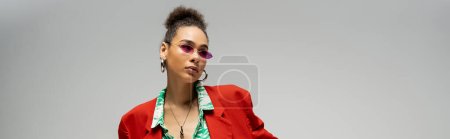 stylowa Afroamerykanka w różowych okularach przeciwsłonecznych i modnym, tętniącym życiem stroju pozowanie na szary, sztandar