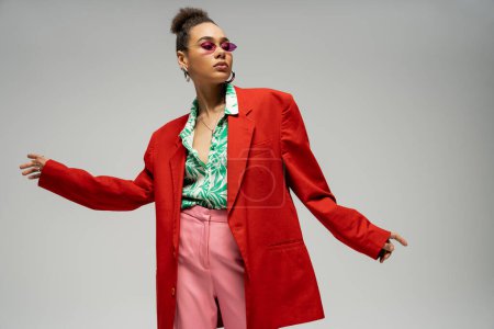 selbstbewusste Afroamerikanerin mit pinkfarbener Brille und trendiger, lebendiger Kleidung posiert vor grauem Hintergrund