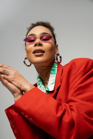 young african american woman in  pink sunglasses and vibrant attire on grey backdrop, accessories