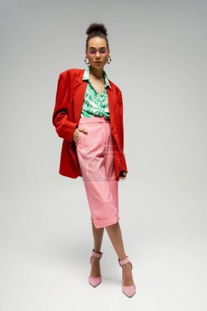 african american woman in  pink glasses and bold style attire posing on grey backdrop, full length