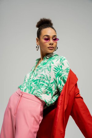 Afrykańska amerykanka w okularach przeciwsłonecznych i modnym stroju pozowanie na szarym tle, styl osobisty