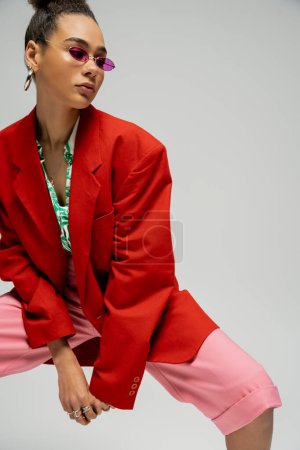 african american woman in  sunglasses and trendy outfit posing on grey backdrop, personal style