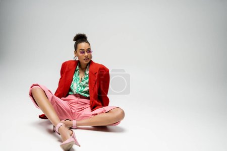 fashionable african american woman in bold style attire and sunglasses sitting on grey backdrop