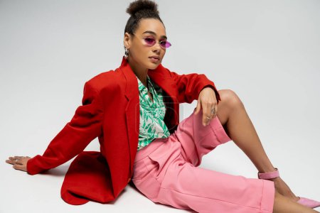 confident african american woman in stylish attire and pink sunglasses sitting on grey backdrop