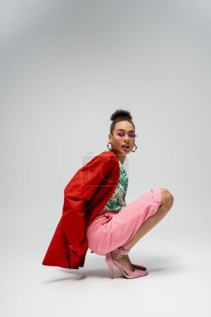 young african american model in stylish attire and pink sunglasses sitting on grey backdrop