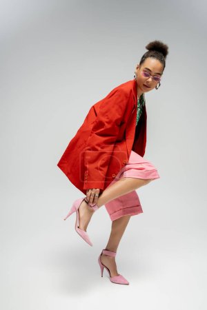 modelo de moda afroamericana en blazer rojo ajustando correa en tacones altos sobre fondo gris