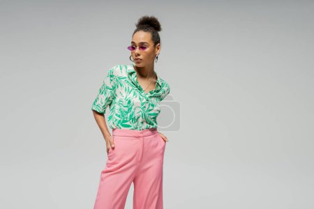 stylish african american woman in pink sunglasses shirt with green pattern posing on grey backdrop