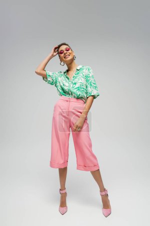 happy african american woman in pink sunglasses and stylish attire posing on grey backdrop