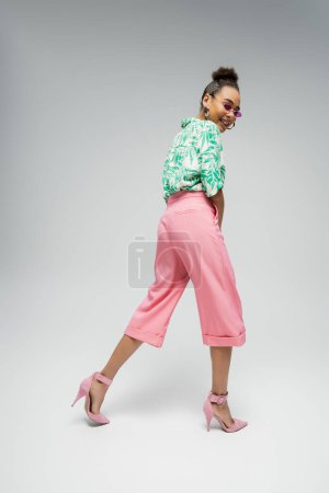 mujer afroamericana feliz en gafas de sol de color rosa y vestimenta elegante caminando sobre fondo gris