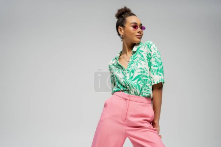 pleased african american woman in pink sunglasses and stylish attire posing on grey backdrop