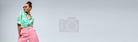 Téléchargez les photos : Femme afro-américaine heureuse en lunettes de soleil roses et tenue élégante posant sur fond gris, bannière - en image libre de droit