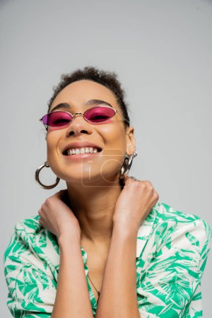Téléchargez les photos : Femme afro-américaine gaie en lunettes de soleil roses et tenue élégante posant sur fond gris - en image libre de droit