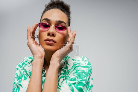 curly african american woman wearing pink sunglasses and posing in stylish attire on grey backdrop