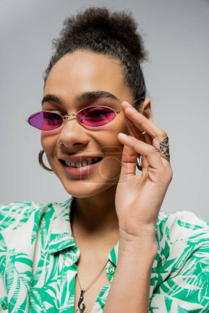 retrato de una mujer afroamericana positiva con gafas de sol rosas y sonriendo sobre un fondo gris