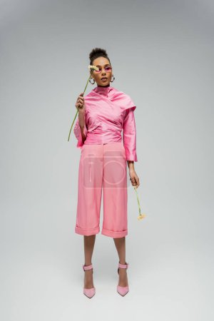 full length of stylish african american woman in pink attire and sunglasses posing with flowers