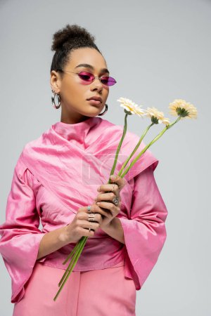 Photo for Stylish african american woman in pink attire and sunglasses posing with flowers on grey backdrop - Royalty Free Image