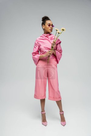 Afrikanische Amerikanerin in rosa Outfit und High Heels posiert mit Blumen vor grauem Hintergrund