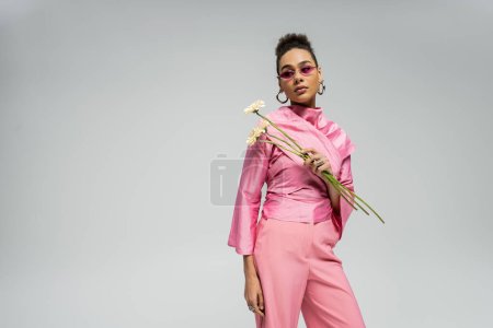 elegant african american woman in pink outfit and sunglasses posing with flowers on grey backdrop