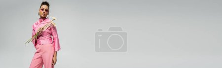 Téléchargez les photos : Élégante femme afro-américaine en tenue rose et lunettes de soleil posant avec des fleurs sur gris, bannière - en image libre de droit