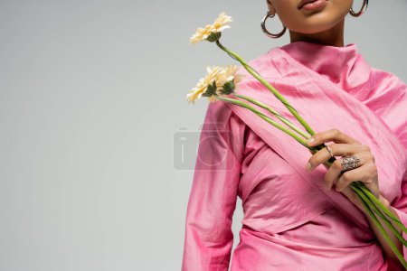mujer afroamericana recortada en traje rosa posando con flores sobre fondo gris, delicado