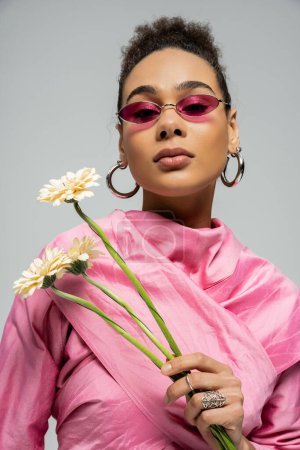 trendiges afrikanisch-amerikanisches Model in rosa Outfit und Sonnenbrille posiert mit Blumen auf grau