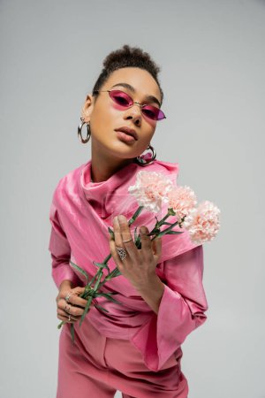 Ausdrucksstarkes afrikanisch-amerikanisches Model in pinkfarbener Kleidung und Sonnenbrille posiert mit Blumen auf grau