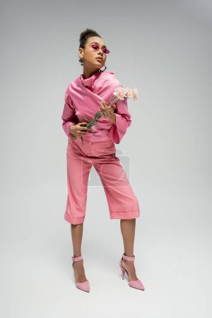 expressive african american fashion model in pink attire and heels posing with flowers, full length