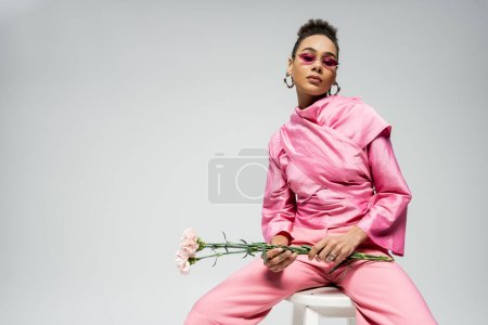 modelo afroamericano de moda en traje rosa y gafas de sol sosteniendo flores y sentado en la silla