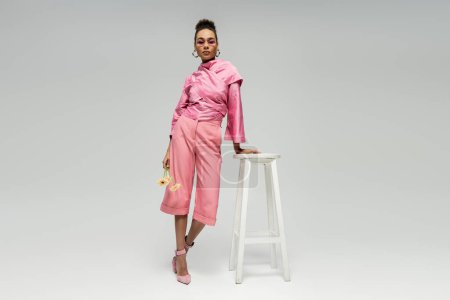 african american fashion model in pink attire and sunglasses posing with flowers near stool on grey