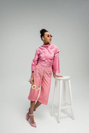 african american young woman in pink attire and sunglasses posing with flowers near stool on grey