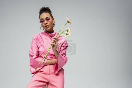 african american young woman in pink attire and sunglasses posing with flowers on grey backdrop