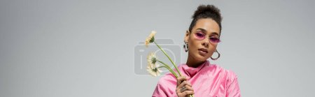 Téléchargez les photos : Jeune femme afro-américaine en tenue rose et lunettes de soleil posant avec des fleurs sur gris, bannière - en image libre de droit