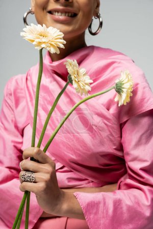 Foto de Vista recortada de la alegre mujer afroamericana en traje rosa sosteniendo flores sobre fondo gris - Imagen libre de derechos