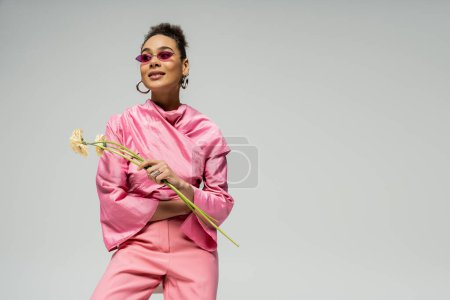 Photo for Happy african american woman in pink attire and sunglasses holding flowers on grey backdrop - Royalty Free Image