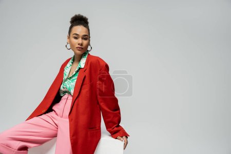 stylisches afrikanisch-amerikanisches Model im trendigen und lebendigen Outfit sitzt auf einem Würfel vor grauem Hintergrund