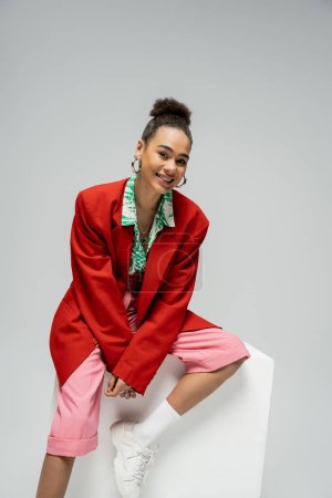 cheerful african american model in trendy and vibrant outfit sitting on cube on grey backdrop